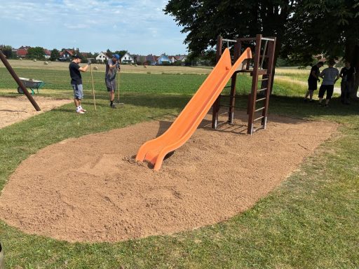 Rutschbahn aus orangefarbenem Kunststoff auf einem Sandspielplatz in einer Wiesenlandschaft.
